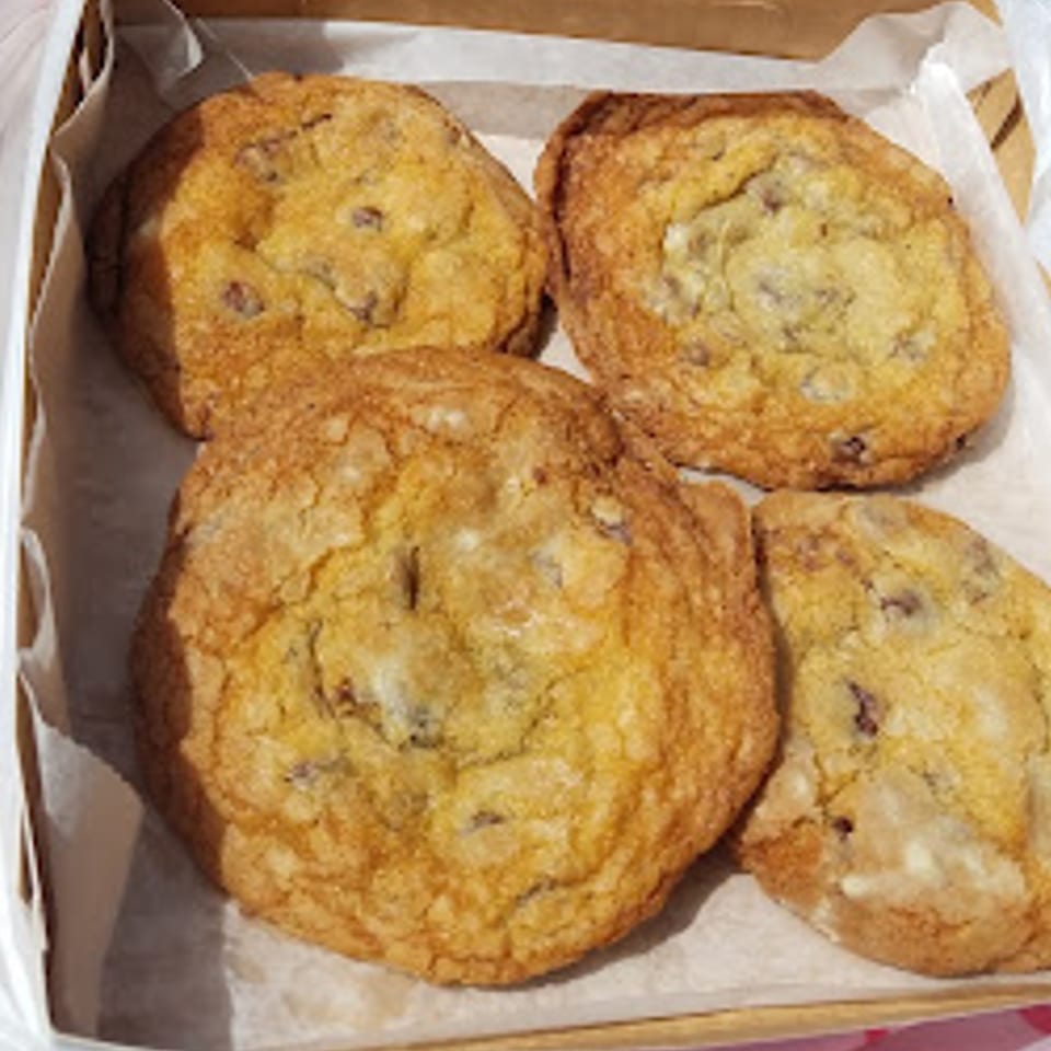 Four golden-brown chocolate chip cookies from The Dough Co. in a white paper-lined box.
