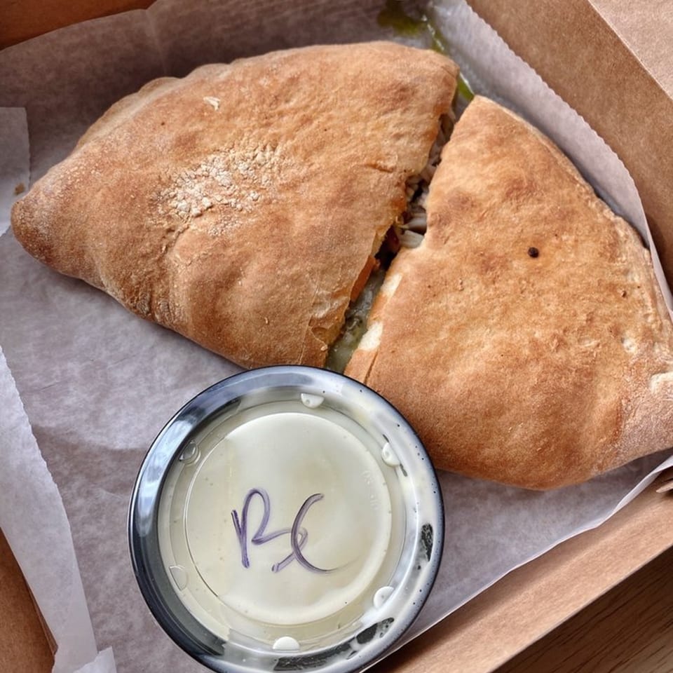 A calzone from The Dough Co. cut in half, with a side of dipping sauce in a take-out box.