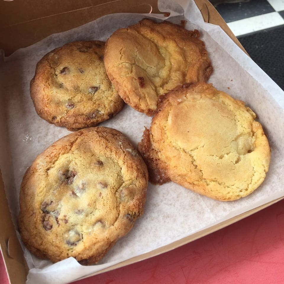 Four warm, golden-brown cookies, two with chocolate chips, in a paper-lined box from The Dough Co.